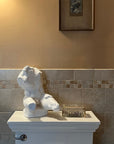image of white plaster sculpture of nude male torso atop white toilet with gray and orange-brown tiling and orange-brown wall behind