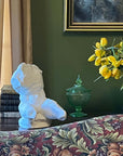 image of white plaster sculpture of nude male torso on brown sofa table behind red and green floral-patterned sofa next to antique books and black vase with yellow tulips with green wall and tan curtains behind