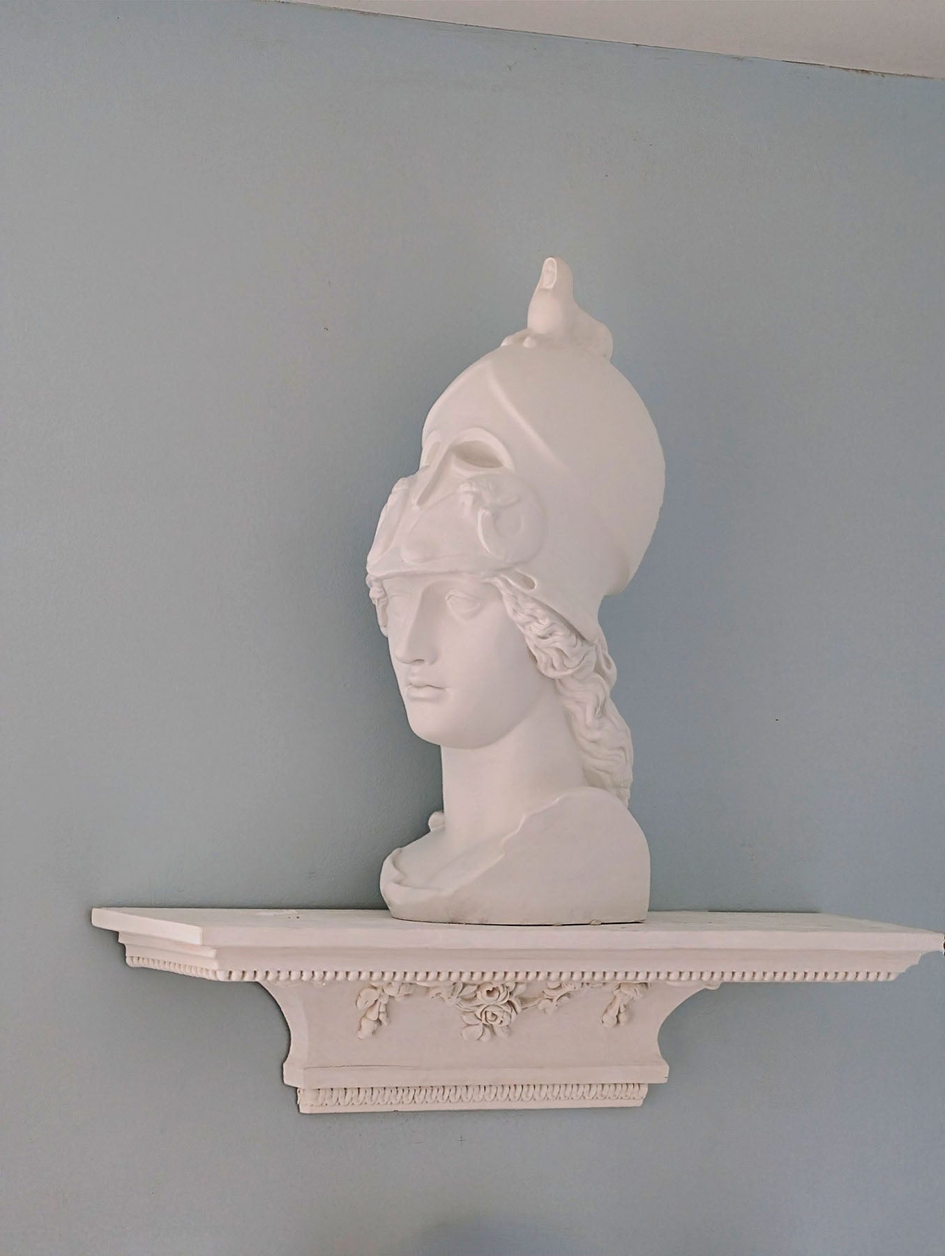 photo of white plaster cast sculpture bust of female head, namely the goddess Athena, wearing a helmet and placed on a matching white ornamental floating shelf on light blue wall