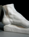 photo of white plaster cast sculpture of left foot on base with green and black books leaning against it against gray and black background
