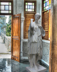 photo of white plaster cast of female figure, namely the goddess Diana, in robes about to clasp two pieces together at proper right shoulder, in corner of room with green marble floor, wooden doors and window treatments, and wall ornamentation, door open to outside stone terrace