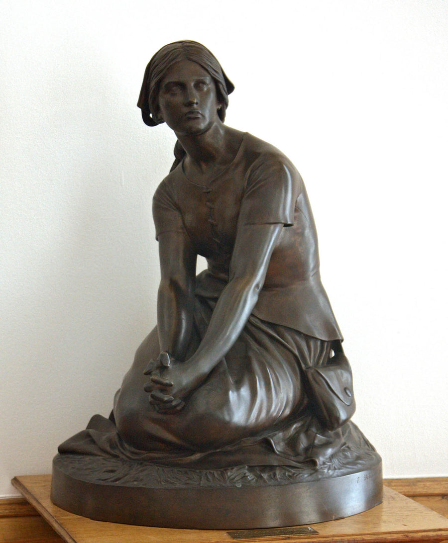 photo of bronze-colored plaster cast sculpture of female, namely Joan of Arc, kneeling with hands on lap on a wood pedestal with white wall behind