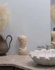 Photo of Plaster Cast sculpture bust of goddess Hebe on neutral bathroom sink with raised white flower basin and brown-bronze pitcher with flowers against light blue wall