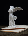 photo of white plaster cast sculpture of winged, headless female figure with flowing drapery next to a pencil atop an open notebook on a wood surface against a gray background