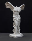 photo of white plaster cast sculpture of winged, headless female figure with flowing drapery on a gray background
