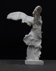 back view photo of white plaster cast sculpture of winged, headless female figure with flowing drapery on a gray background