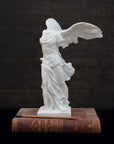 photo of white plaster cast sculpture of winged, headless female figure with flowing drapery next to a pencil atop an antique red book against a dark gray wood background