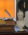 plaster cast bust sculpture of man with beard, namely Lincoln, on antique wooden table decorated with antique books, candles, and other paraphernalia and two wood and wicker chairs beside the table and sunlight coming in shaded window behind