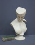 photo of off-white plaster cast sculpture bust of female without top of head and with piece of drapery on socle base with green book laying beside it on gray background