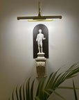 photo of plaster cast sculpture of classical male figure, namely Hercules, wall-mounted atop a plaster cast bracket with leaf design and top lit for nighttime viewing on a neutral wall and plant leaves visible in foreground