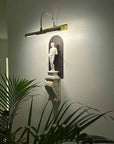 photo of plaster cast sculpture of classical male figure, namely Hercules, wall-mounted atop a plaster cast bracket with leaf design and top lit for nighttime viewing on a neutral wall and plant leaves visible in foreground