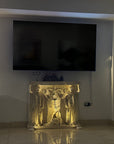 photo of plaster cast of free-standing relief sculpture with three female figures, two of which are caryatides, against a living room wall with a television mounted above it and wooden desk and other decor nearby, and relief floor lit for nighttime viewing