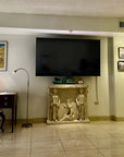 photo of plaster cast of free-standing relief sculpture with three female figures, two of which are caryatides, against a living room wall with a television mounted above it and wooden desk and other decor nearby