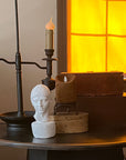 photo of white plaster cast head of male on small round table decorated with wood boxes and pillar candle and candelabra with sunlight coming in shaded window behind