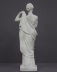 photo of white plaster cast statue of female in robes with left arm raised and holding robe on gray background