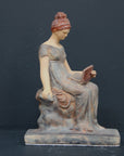 Photo of plaster cast sculpture of female in green-brown tinted robes seated with opened book against grey background