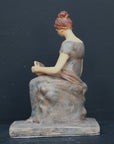 Photo of plaster cast sculpture of female in green-brown tinted robes seated with opened book against grey background