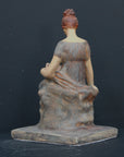 Photo of plaster cast sculpture of female in green-brown tinted robes seated with opened book against grey background