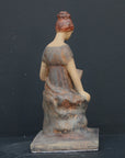 Photo of plaster cast sculpture of female in green-brown tinted robes seated with opened book against grey background
