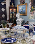 photo of plaster cast sculpture bust of man, namely the god Apollo, with hair piled high in the front and a broach near his neck holding robes and set on a wooden table in the background against a blue pinstriped wall and surrounded by antiques including a table in the foreground with white and blue china and crystal