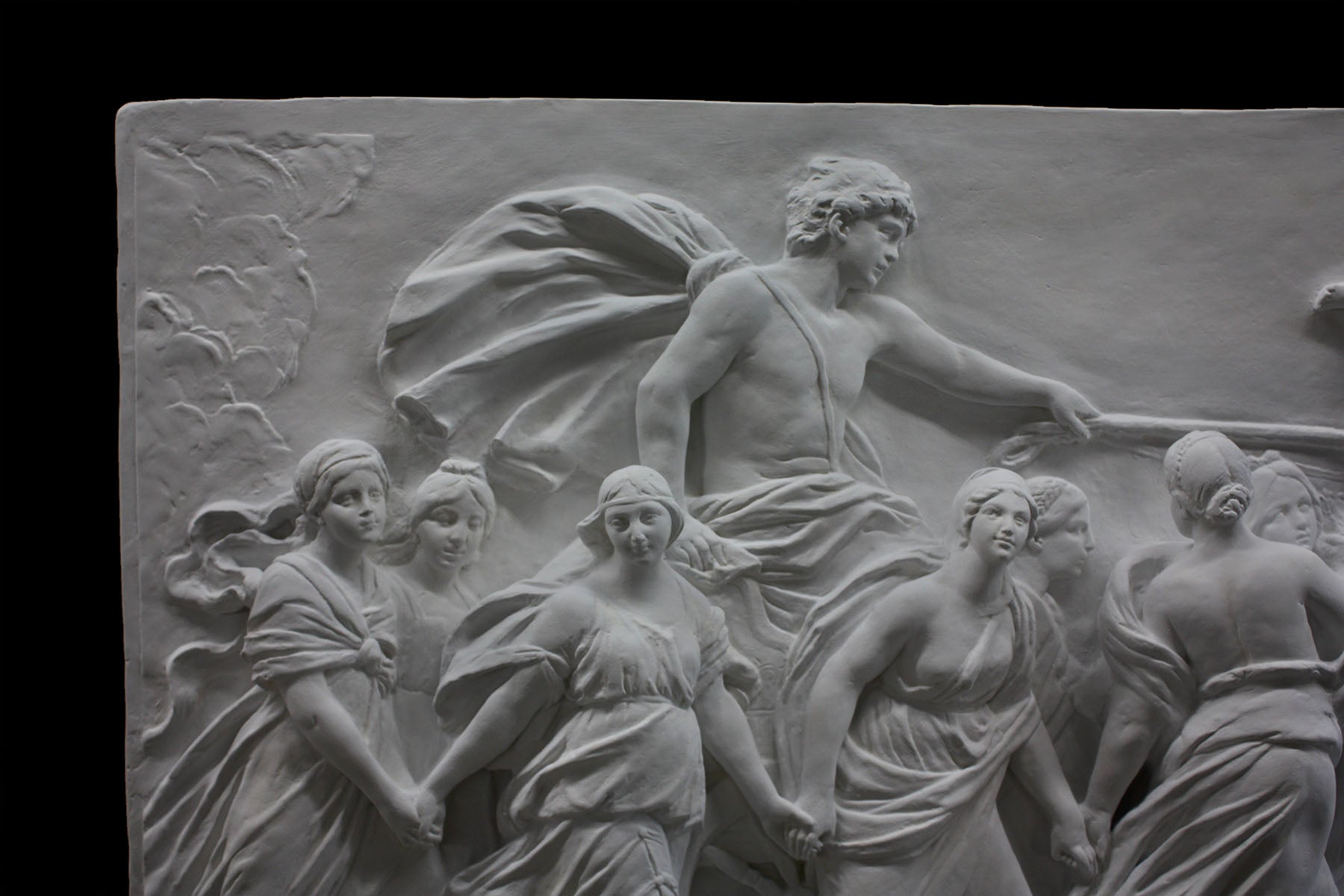 detail photo of a plaster cast sculpture relief of a woman, namely the goddess Aurora, flying and leading a chariot with a man pulled by horses towards the right while a cherub and other women fill the scene, on a black background