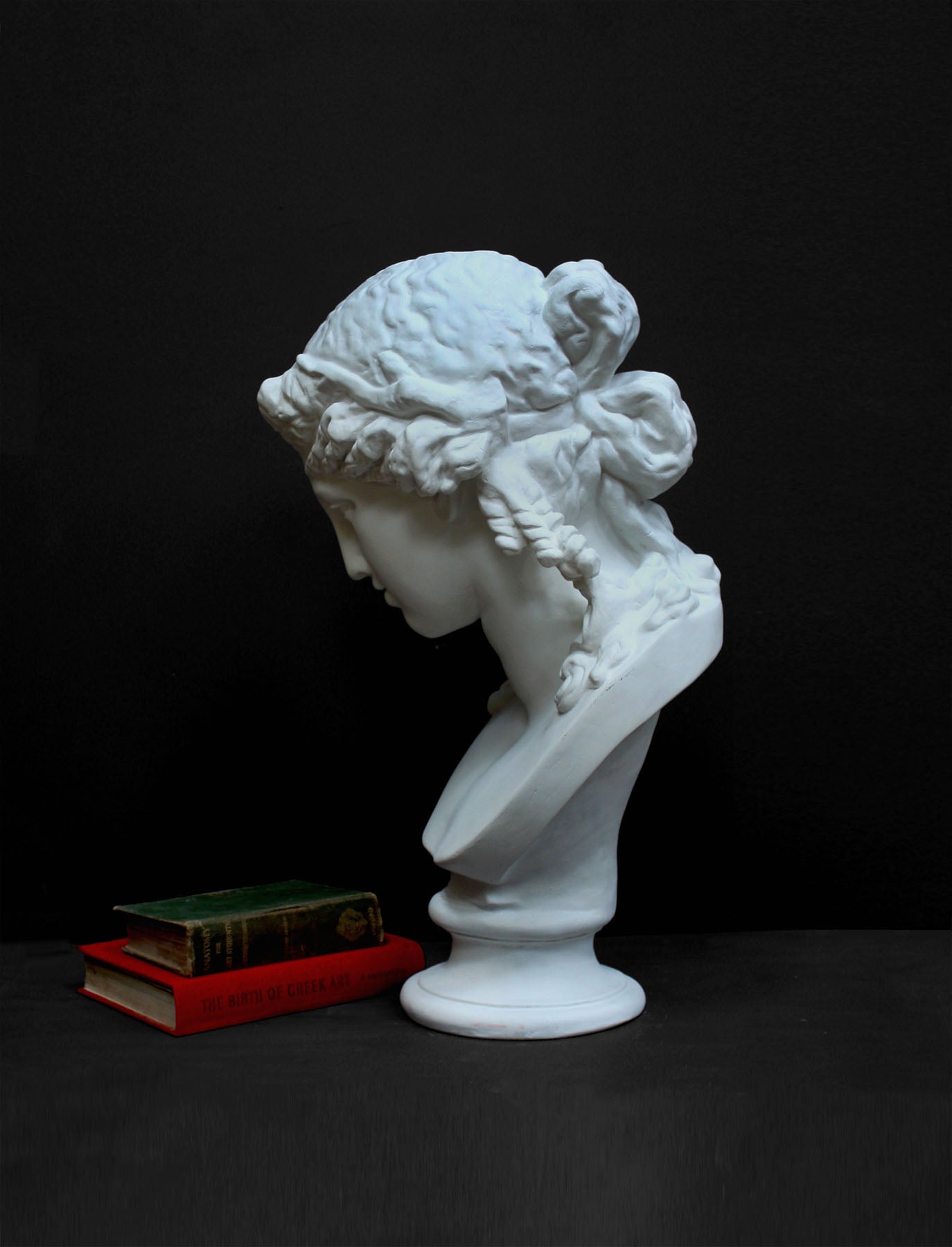 photo of plaster cast of sculpture bust of Ariadne or Bacchus with long, curly hair and head band next to books on a gray background
