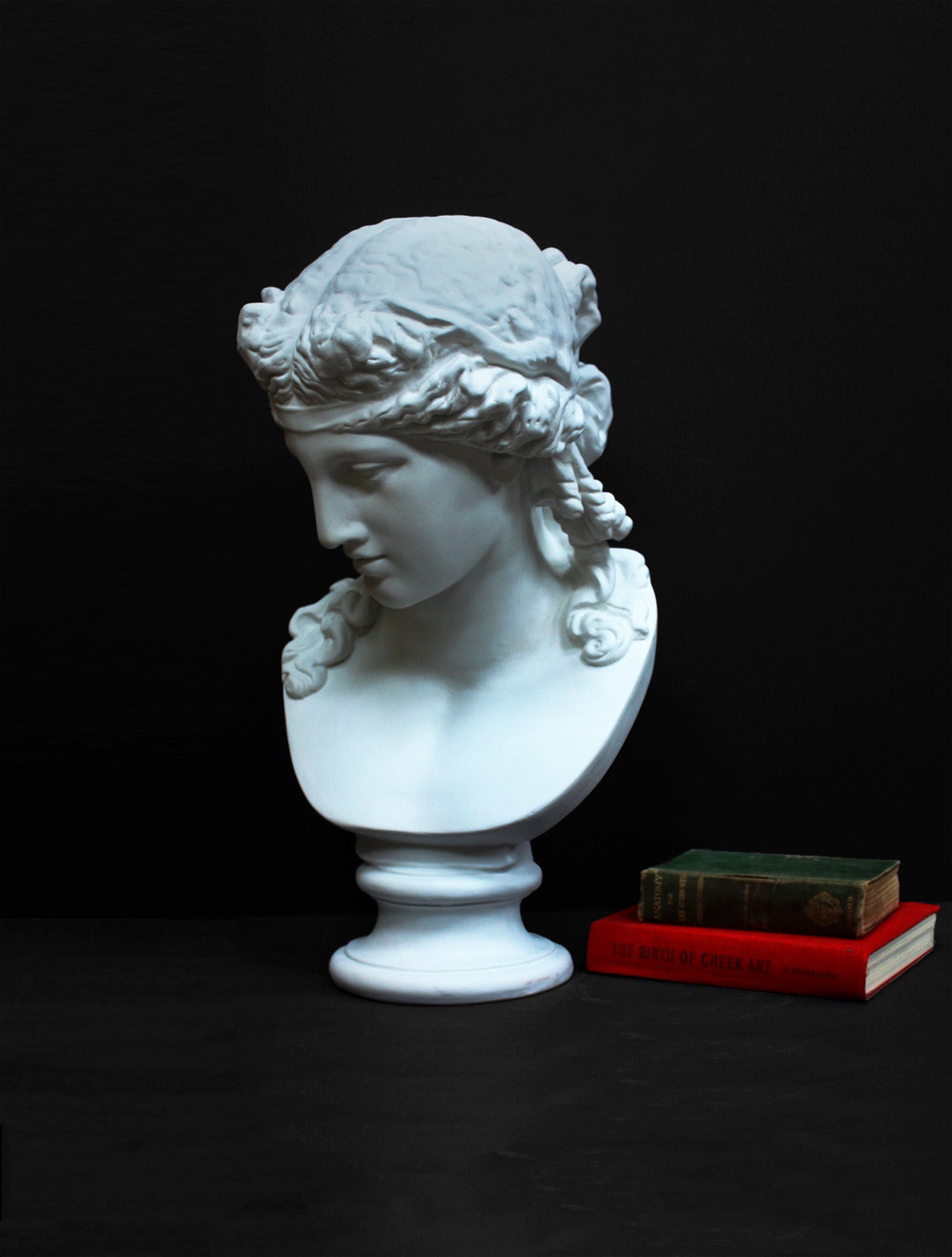 photo of plaster cast of sculpture bust of Ariadne or Bacchus with long, curly hair and head band next to books on a gray background