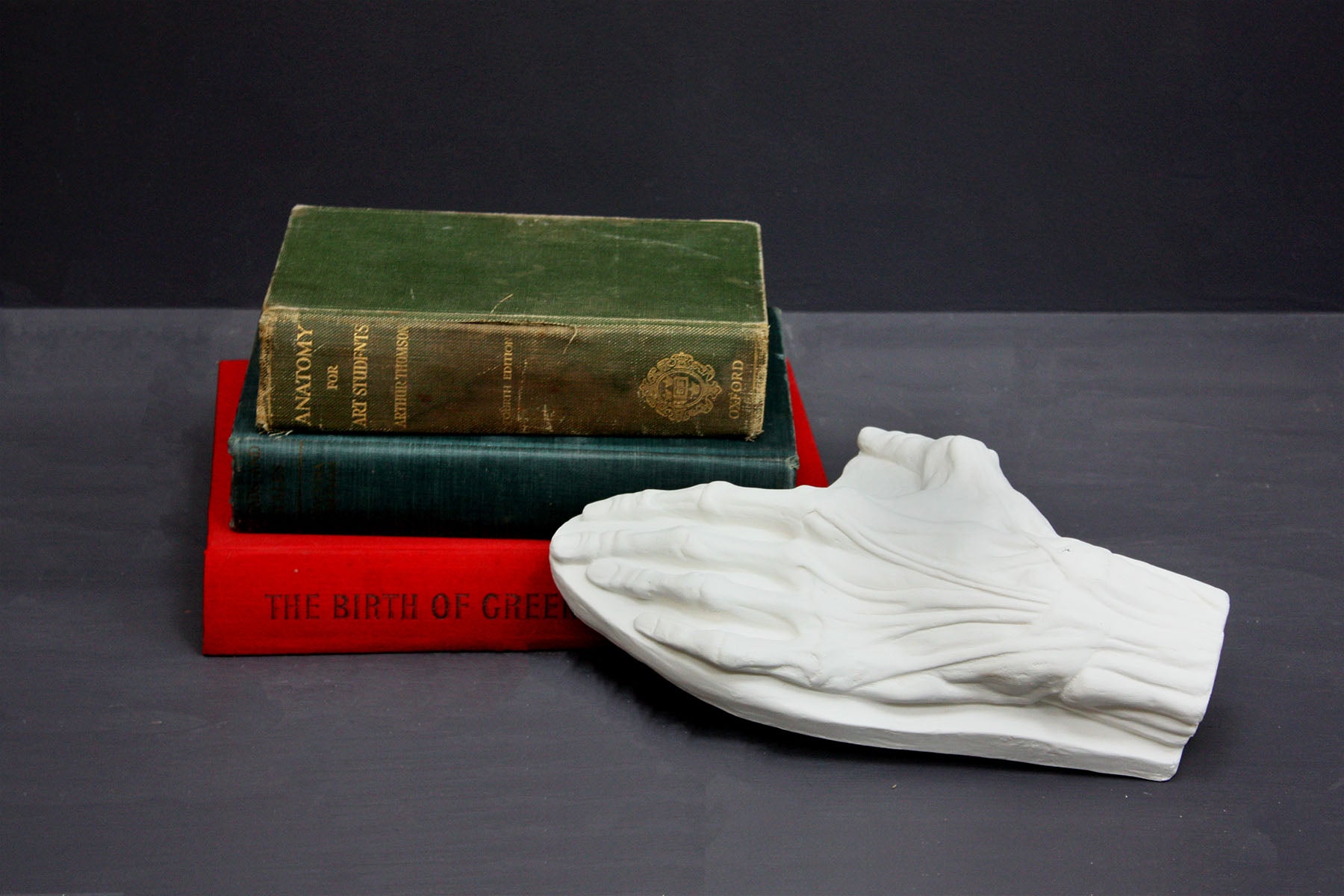 Photo of a plaster cast of a flayed hand for artist study next to three books on a gray background