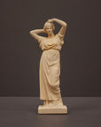 photo of off-white plaster cast statue of a female in robes with her arms raised to her head against gray background