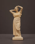 photo of off-white plaster cast statue of a female in robes with her arms raised to her head against gray background
