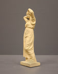 photo of off-white plaster cast statue of a female in robes with her arms raised to her head against gray background