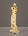 photo of off-white plaster cast statue of a female in robes with her arms raised to her head against gray background