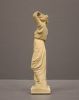 photo of off-white plaster cast statue of a female in robes with her arms raised to her head against gray background