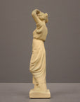 photo of off-white plaster cast statue of a female in robes with her arms raised to her head against gray background