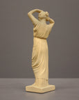 photo of off-white plaster cast statue of a female in robes with her arms raised to her head against gray background