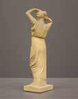 photo of off-white plaster cast statue of a female in robes with her arms raised to her head against gray background