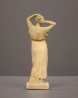photo of off-white plaster cast statue of a female in robes with her arms raised to her head against gray background