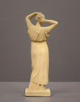 photo of off-white plaster cast statue of a female in robes with her arms raised to her head against gray background