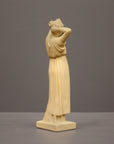 photo of off-white plaster cast statue of a female in robes with her arms raised to her head against gray background