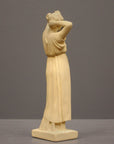 photo of off-white plaster cast statue of a female in robes with her arms raised to her head against gray background