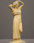 photo of off-white plaster cast statue of a female in robes with her arms raised to her head against gray background