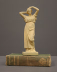 photo of off-white plaster cast statue of a female in robes with her arms raised to her head atop green book against gray background