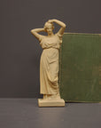 photo of off-white plaster cast statue of a female in robes with her arms raised to her head beside green book against gray background