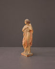 photo of plaster cast statue of female in pink robes with left arm raised and holding robe on gray background