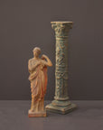 photo of plaster cast statue of female in pink robes with left arm raised and holding robe beside a column-styled candlestick on gray background
