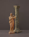 photo of plaster cast statue of female in pink robes with left arm raised and holding robe beside a column-styled candlestick on gray background