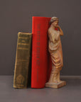 photo of plaster cast statue of female in pink robes with left arm raised and holding robe beside two books on gray background