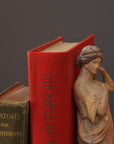 photo of plaster cast statue of female in pink robes with left arm raised and holding robe beside two books on gray background