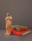 photo of plaster cast statue of female in pink robes with left arm raised and holding robe beside two stacked books on gray background