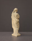 photo of off-white plaster cast statue of female in robes with left arm raised and holding robe on gray background